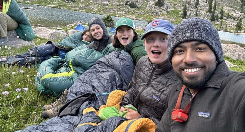 a group of people lay or sit on the ground, bundled in coats and sleeping backs and smile for the photo 
